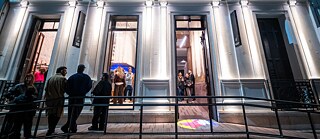 Vista nocturna exterior del edificio histórico iluminado con fachada clásica del Kultur Ensemble Córdoba; varias personas entran y salen del acceso.