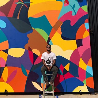 De Leon Peres (Jotape Pax) está sentado em um banco alto em frente a um grande mural colorido com formas abstratas e perfis de rostos. De Leon Peres usa camiseta clara, óculos escuros e calça escura, mantendo as mãos apoiadas entre as pernas. O fundo é dominado por cores vibrantes como amarelo, azul, laranja e rosa, criando um contraste forte com a figura central.