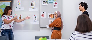Eine Lehrerin erklärt in einem Deutschkurs Lebensmittelvokabeln an der Tafel, während mehrere Lernende aufmerksam zuhören.