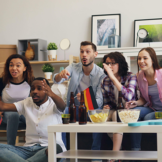 A group of people sitting on a couch watching TV with a German flag. Popcorn and chips are on the TV table.