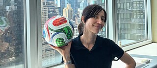 Lisa Hollenbach, deren schwarzes Haar zu einem Pferdeschwanz zusammengebunden ist, hält den offiziellen WM-Fußball in der Hand und steht vor dem Fenster des Generalkonsulats der Bundesrepublik Deutschland in New York, im Hintergrund sind Wolkenkratzer zu sehen.
