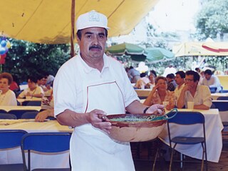 Un hombre con una gorra del aniversario y un bol de salsa verde en sus manos.