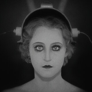 Imagen en blanco y negro con estética de cine temprano: primer plano de un rostro de mujer con ojos grandes y muy resaltados. Sobre la cabeza hay un dispositivo técnico similar a un casco con piezas laterales. El fondo muestra cortinas oscuras.