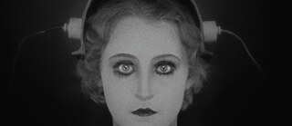 Imagen en blanco y negro con estética de cine temprano: primer plano de un rostro de mujer con ojos grandes y muy resaltados. Sobre la cabeza hay un dispositivo técnico similar a un casco con piezas laterales. El fondo muestra cortinas oscuras.