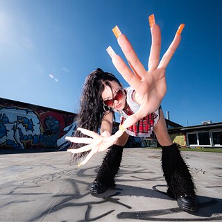 Portrait of Mariybu with black ponytail and sunglasses bending down towards the camera and reaching out her hand with long fingernails
