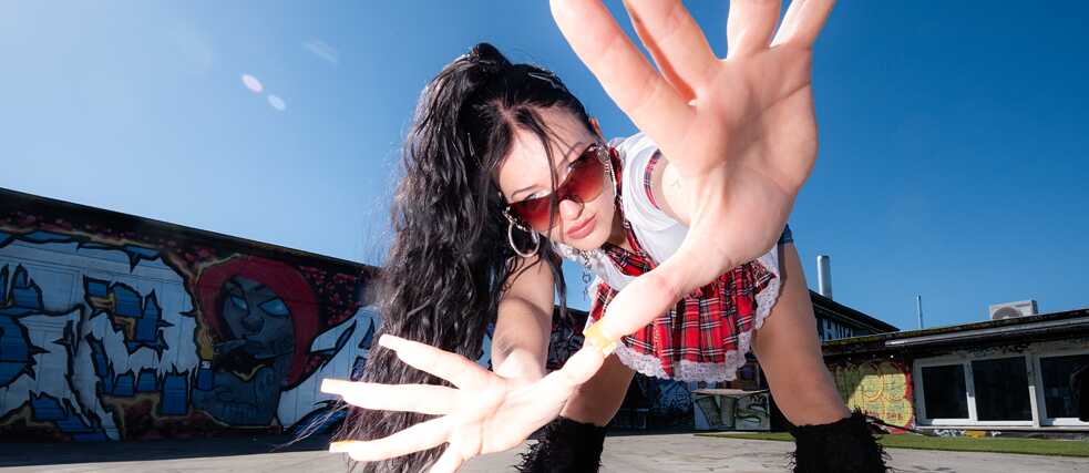 Artist Mariybu, black hair in a ponytail and sunglasses,  bending down and holding a hand with long fingernails close to the camera 