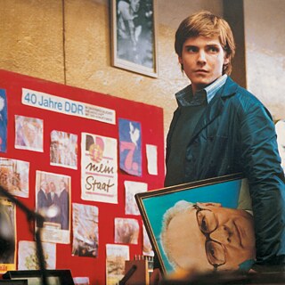 Alex Kerner (Daniel Brühl) hält das Porträt von Erich Honecker, dem Staatschef der DDR, vor einer „40 Jahre DDR“-Tafel in einem Innenraum mit DDR-Bildern