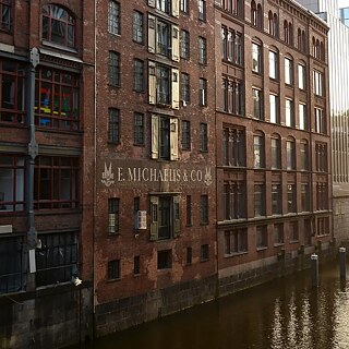 Historisches Backsteingebäude des Westwerks in Hamburg am Ufer eines Kanals in der Speicherstadt. Die mehrstöckige Fassade mit roten Ziegeln, großen Sprossenfenstern und verblasster Aufschrift „E. Michaelis & Co.“ wird von warmem Sonnenlicht beleuchtet. Rechts grenzen moderne Glas- und Bürogebäude an das historische Ensemble. Im Vordergrund verläuft ein Wasserkanal mit Dalben und einer kleinen Brücke beziehungsweise Unterführung. Die Szene zeigt den Kontrast zwischen historischer Industriearchitektur und moderner Stadtentwicklung.