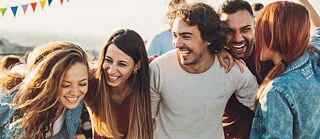 Mehrere Personen stehen eng beieinander im Freien und blicken nach unten oder zur Seite. Die Szene wirkt ungezwungen und zeigt eine gemeinsame Aktivität bei warmem Tageslicht.