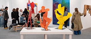 Visitors at a contemporary art fair viewing colorful sculptural works and paintings in a bright gallery space.