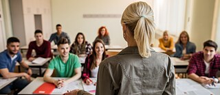 Rückansicht auf eine Lehrerin, die zu Schülern der Sekundarstufe im Klassenraum spricht.