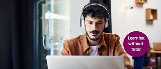 A man sits facing a laptop, wearing headphones and looking focused at the screen while studying German through self-paced online learning.
