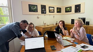 Vier Teilnehmende arbeiten mit Laptops und Unterlagen, während eine Person sich über den Tisch beugt und die Gruppe bei einer gemeinsamen Aufgabe unterstützt.