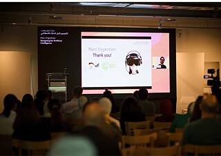 Blick auf das Publikum während einer Präsentation bei einer Designkonferenz. Auf der Leinwand sind Vortragsfolien zum Thema künstliche Intelligenz zu sehen.