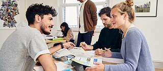 Mehrere Erwachsene sitzen an einem Tisch in einem hellen Unterrichts- oder Seminarraum, lesen Unterlagen und besprechen Inhalte. Im Hintergrund arbeiten weitere Personen an Schreibtischen vor großen Fenstern.