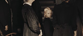 Ein blondes Mädchen mit geflochtenen Zöpfen steht inmitten dunkel gekleideter Erwachsener und blickt direkt in die Kamera.