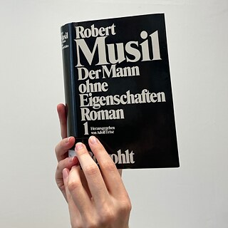 Eine Hand hält den Roman Der Mann ohne Eigenschaften von Robert Musil vor hellem Hintergrund. Das schwarze Buchcover mit großer weißer Typografie steht im Mittelpunkt und zeigt deutlich Titel, Autor und Verlag.
