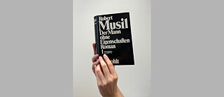 Eine Hand hält den Roman Der Mann ohne Eigenschaften von Robert Musil vor hellem Hintergrund. Das schwarze Buchcover mit großer weißer Typografie steht im Mittelpunkt und zeigt deutlich Titel, Autor und Verlag.