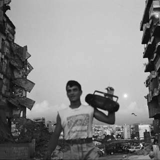 Schwarz-Weiß-Aufnahme eines jungen Mannes, der mit einem Ghettoblaster auf der Schulter durch eine von zerstörten Gebäuden gesäumte Straße geht.