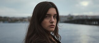 Porträtaufnahme einer jungen Frau mit langen braunen Haaren und einer kleinen Verletzung an der Augenbraue, die vor einer Wasserkulisse direkt in die Kamera blickt.