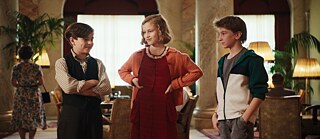 Tres niños se encuentran de pie en un amplio salón con columnas, mesas, sillones y lámparas de pie. Una persona viste un vestido rojo con cárdigan, mientras que las otras dos llevan ropa informal y de estilo clásico. Al fondo se observan otras personas y elementos del interior del edificio.