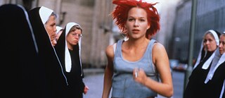 Joven mujer de cabello rojo intenso corre por una calle estrecha de la ciudad. A ambos lados hay varias personas con hábitos negros y cofias blancas. Al fondo se observan edificios antiguos y una escena urbana ligeramente desenfocada.