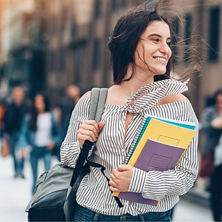 Una mujer joven con mochila lleva varios cuadernos y carpetas mientras camina por una calle concurrida. La imagen muestra a la persona en primer plano, con transeúntes y edificios desenfocados al fondo. La fotografía fue tomada al aire libre con luz diurna.