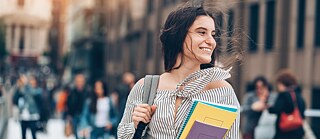 Una mujer joven con mochila lleva varios cuadernos y carpetas mientras camina por una calle concurrida. La imagen muestra a la persona en primer plano, con transeúntes y edificios desenfocados al fondo. La fotografía fue tomada al aire libre con luz diurna.