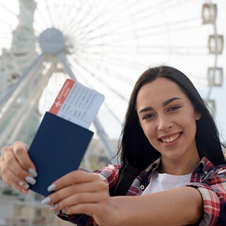 Una mujer sostiene un pasaporte con una tarjeta de embarque delante de la cámara. Al fondo se observa una gran rueda de observación. La fotografía fue tomada al aire libre con luz diurna y muestra a la persona en primer plano frente a la estructura desenfocada de la rueda.