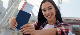 Una mujer sostiene un pasaporte con una tarjeta de embarque delante de la cámara. Al fondo se observa una gran rueda de observación. La fotografía fue tomada al aire libre con luz diurna y muestra a la persona en primer plano frente a la estructura desenfocada de la rueda.