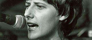 Schwarz-Weiß-Porträt einer Frau mit kurzen Haaren und einem Ansteckbutton auf dem Pullover, die vor einem Mikrofon spricht.