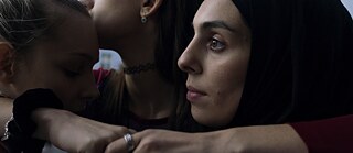 Nahaufnahme von drei jungen Frauen, von denen eine ein schwarzes Kopftuch trägt, die sich in einer engen Umarmung zusammenschließen.