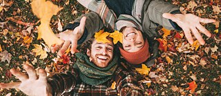 Luftaufnahme von zwei Personen, die auf einer mit bunten Herbstblättern bedeckten Wiese liegen. Mehrere Blätter liegen auf ihrer Kleidung und in ihrer Umgebung, einige werden in die Luft geworfen. Die Hände der Personen sind zur Kamera ausgestreckt. Rechts oben ist ein weißer Kreis mit violetter Schrift zu sehen, auf dem steht: „Date limite CPF : 10/09“.