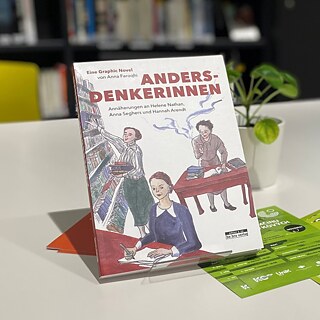 Das Buch „Andersdenkerinnen“ steht auf einem Tisch in einer Bibliothek. Das Cover zeigt gezeichnete Szenen mit drei schreibenden und lesenden Personen. Im Hintergrund sind Bücherregale und eine Zimmerpflanze zu sehen.