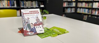 Das Buch „Andersdenkerinnen“ steht auf einem Tisch in einer Bibliothek. Das Cover zeigt gezeichnete Szenen mit drei schreibenden und lesenden Personen. Im Hintergrund sind Bücherregale und eine Zimmerpflanze zu sehen.