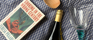 Das Buch 'Cooking in the Wrong Century' liegt mit einer Weinflasche, einem Weinglas und einem Holzkochlöffel auf einer weiß-blaukarierten Tischdecke