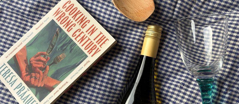 Das Buch 'Cooking in the Wrong Century' liegt mit einer Weinflasche, einem Weinglas und einem Holzkochlöffel auf einer weiß-blaukarierten Tischdecke