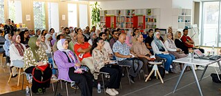  في المكتبة: يجلس عدد كبير من المشاركين في صفوف ويتابعون باهتمام محاضرةً يقدمها ضيف. وتُبرز القاعة المضيئة المزوّدة برفوف الكتب والأجواء التعليمية الطابع الثقافي والتعليمي للفعالية، كما تعكس أهمية الحوار وتبادل المعرفة بين الحاضرين. 