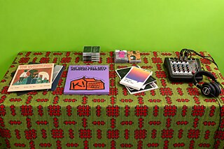 Sonic-Library-Display mit Publikationen, Kassetten, Mischpult und Kopfhörern auf einem gemusterten Tisch.