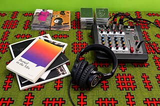 Sonic-Library-Setup mit Kopfhörern, Audiomischpult, CDs und Publikationen auf einem gemusterten Tisch.