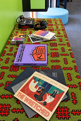 Sonic-Library-Display mit Publikationen, Kassetten, Mischpult und Kopfhörern auf einem gemusterten Tisch.