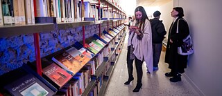 Besucher*innen in einer Bibliothek vor langen Bücherregalen, eine Person blättert in einem Buch.