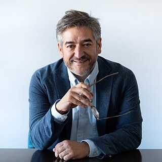 Retrato de Javier Cáceres sentado frente a una mesa, mirando hacia la cámara. Lleva una camisa celeste y un saco azul, y sostiene unos anteojos con una mano. Al fondo se observa una pared clara y lisa.