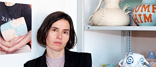 Die Autorin Clara Umbach sitzt neben einem Regal mit Keramikobjekten.