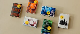 Sechs Bücher der Shortlist des Deutschen Buchpreises 2026 liegen auf einer hellen Oberfläche und sind aus der Vogelperspektive fotografiert.