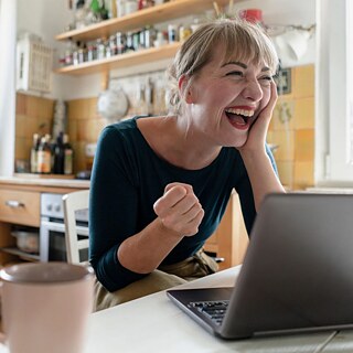 Eine Frau mit Laptop sitzt in ihrer Küche und freut sich über das, was sie am Bildschirm sieht.