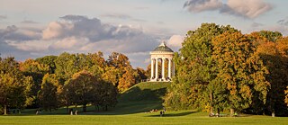 A landscaped park with a folly on a hill, people are strolling along, leaves are turning yellow and brown