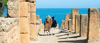 Zwei Frauen, darunter Alice Schwarzer (Autorin), gehen auf einem mit Pfeilern gesäumtem Weg in Richtung Strand von Algerien, das Mittelmeer im Blick.