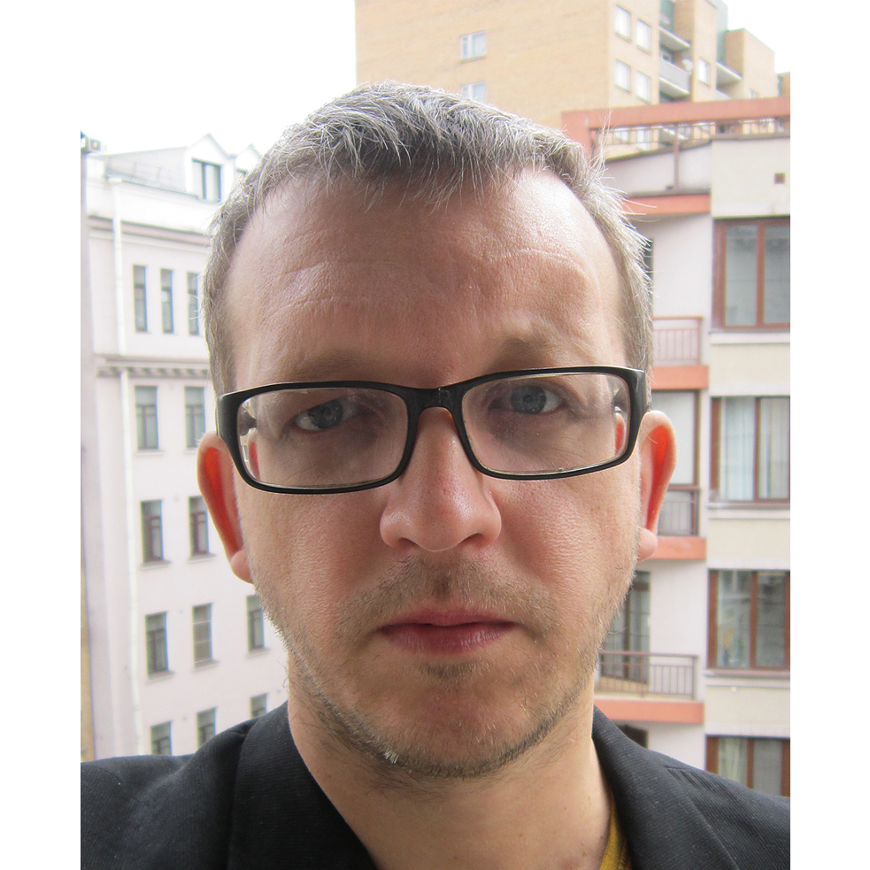 Head shot Oleg Nikiforov; he wears short hair and black rectangular glasses;  in the background buildings