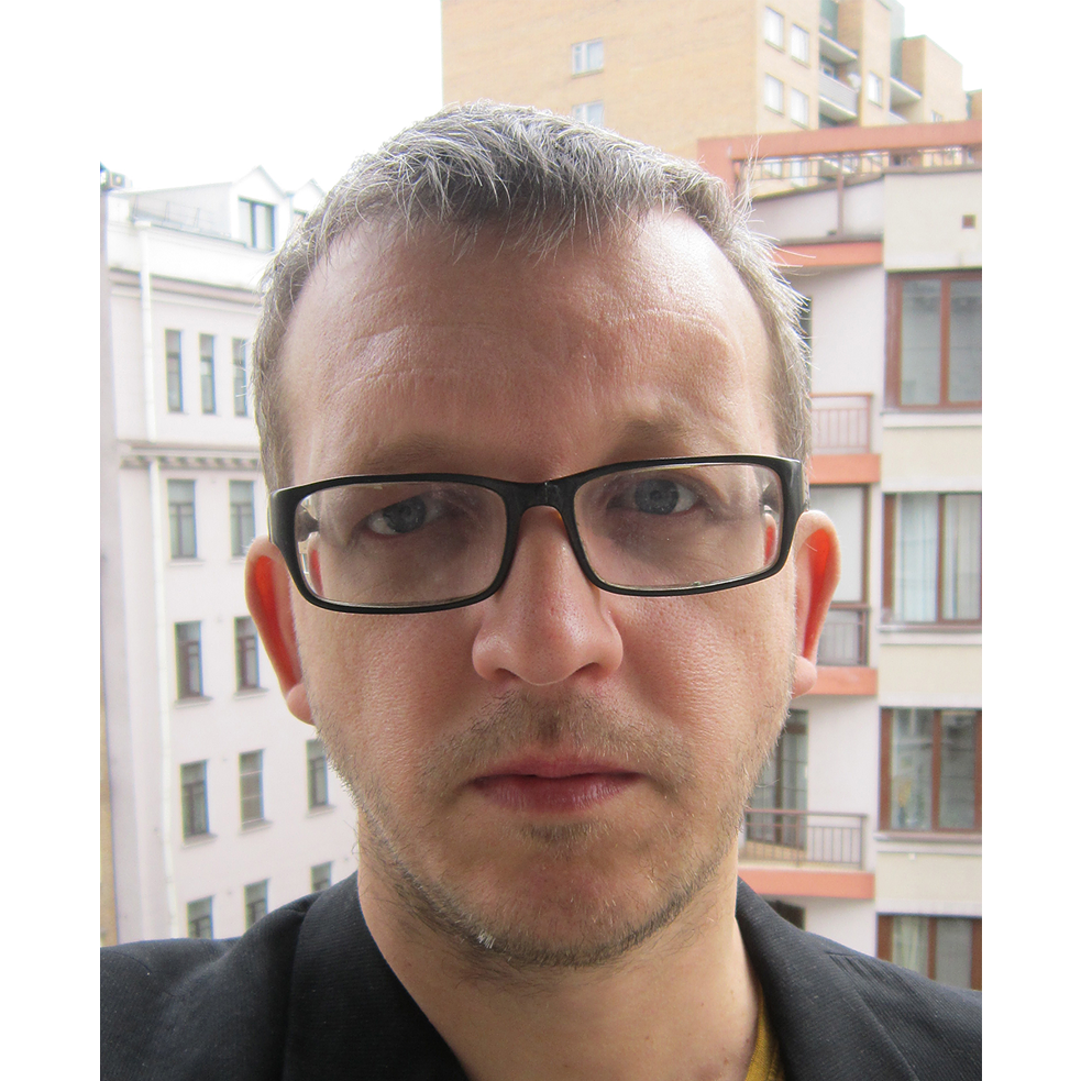 Retrato de Oleg Nikiforov; tiene el pelo corto y usa lentes negros rectangulares; al fondo hay casas.
